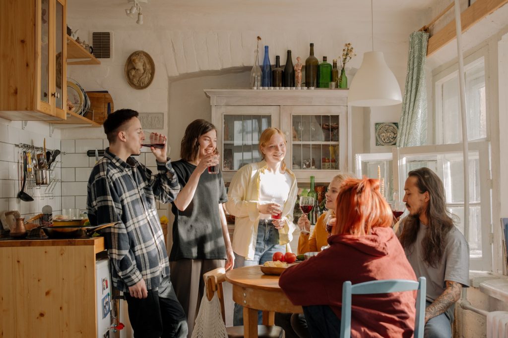 A group of friends gathered around a kitchen table, to socialize. There is a mix of men and women, and most of them are holding glasses in their hands, drinking and smiling to one another.