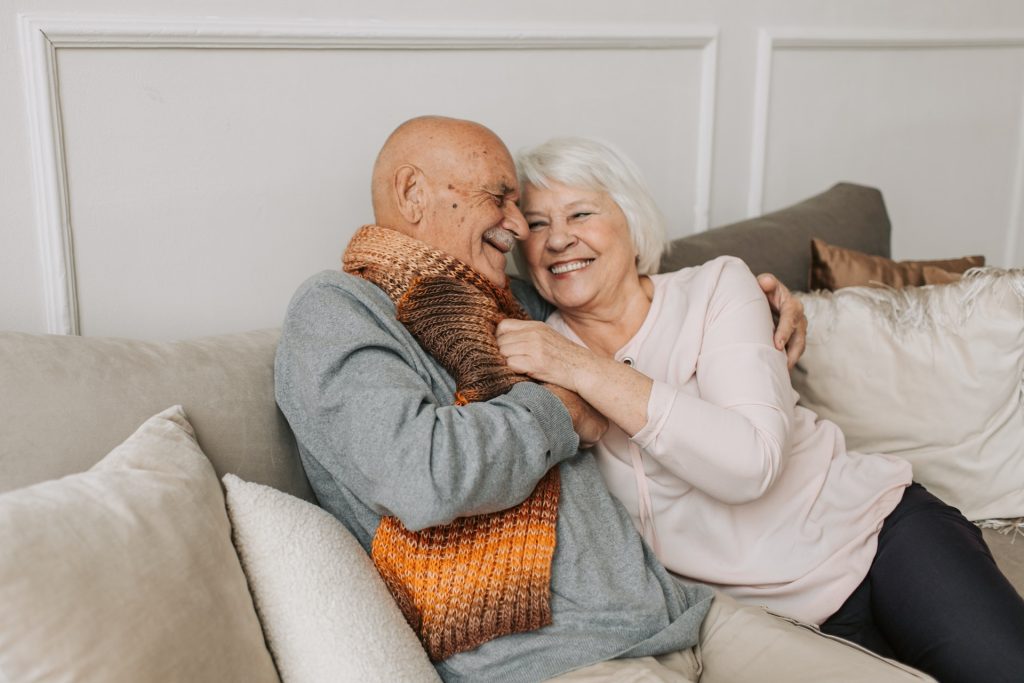 An elderly married couple having a moment of intimacy in the sofa. The man and the woman are smiling and hoding each other affectionately.