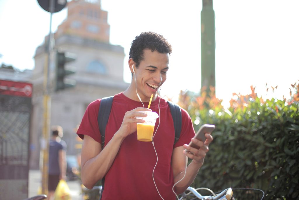 A young man smiling at his phone because he has just received a text from his long-distance girlfriend. He’s also wearing headphones and drinking a glass of orange juice.